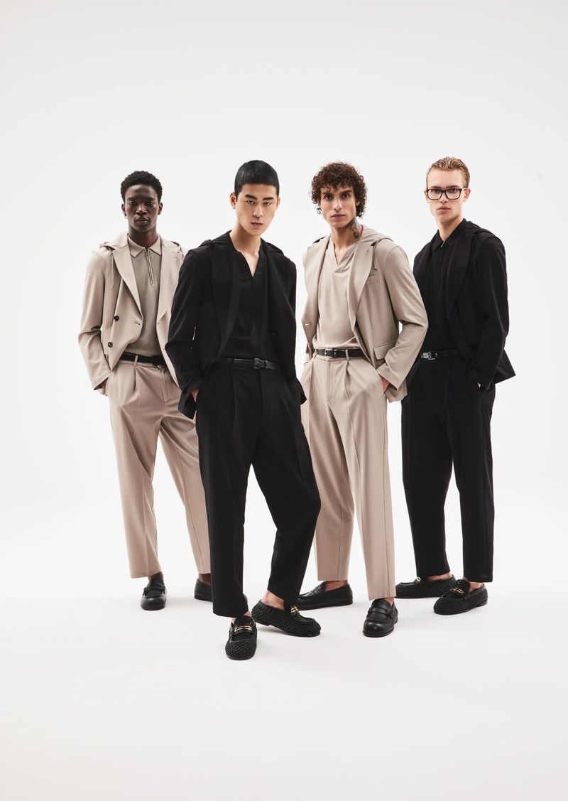Four adult male fashion models pose in neutral and black coloured suits against a bright white backdrop, indoor studio setting.