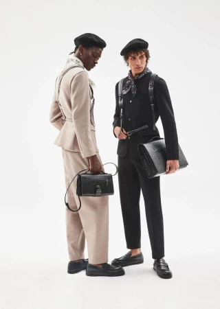 Two male models in berets and tailored suits carrying black leather handbags against a seamless white background.