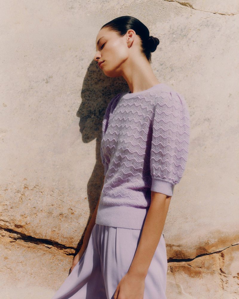 Woman in lavender knit top and pants poses in front of a textured rock wall in natural light, showcasing soft textures and color coordination.