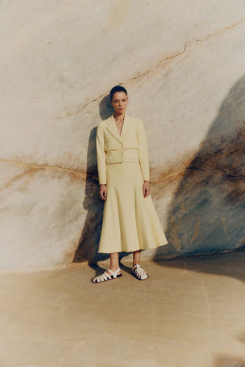 A woman in a light yellow skirt and blazer poses in a photo studio against a stone wall, creating a fashion editorial image.