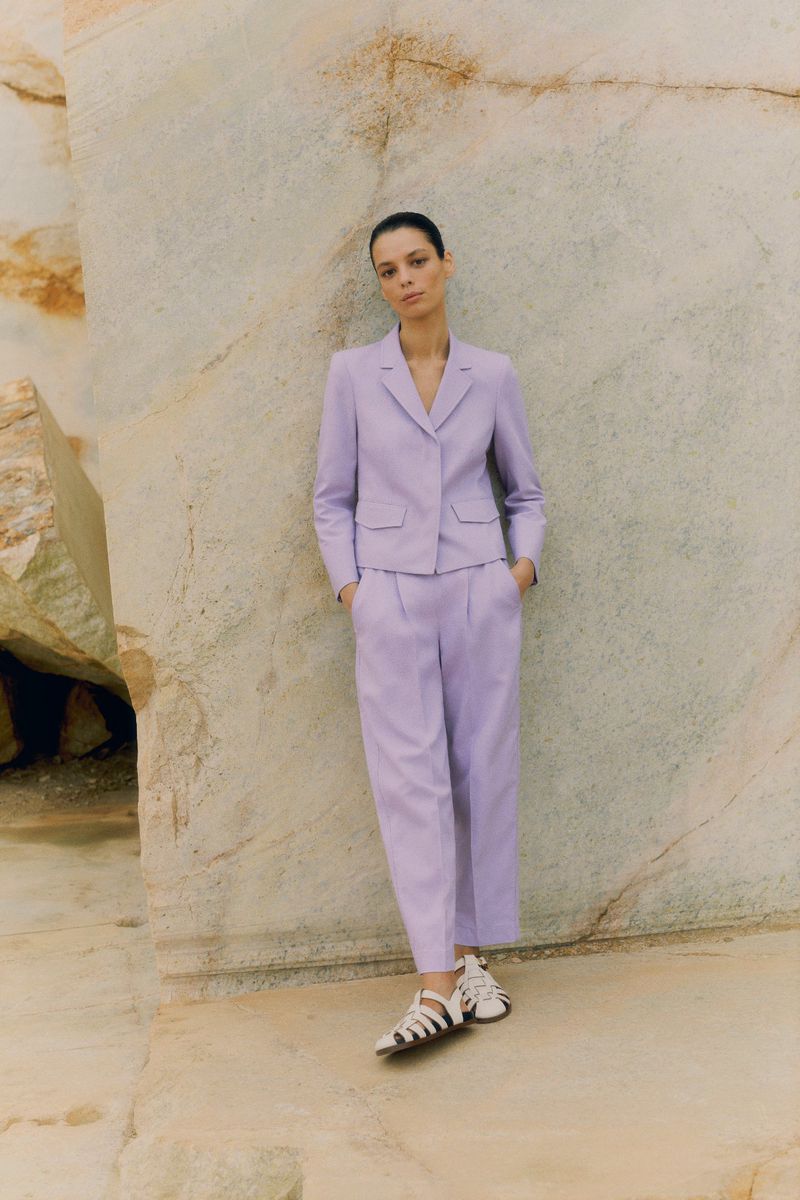 An adult female fashion model poses outdoors wearing a lilac blazer and matching culottes in front of a light-colored rock formation.