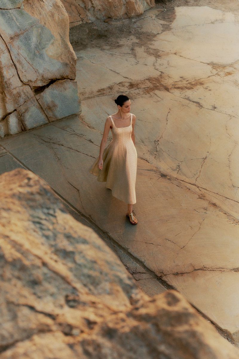 Woman in a beige dress walking on marble rock formation near ocean, bathed in golden light, creating a sense of serenity and natural beauty.
