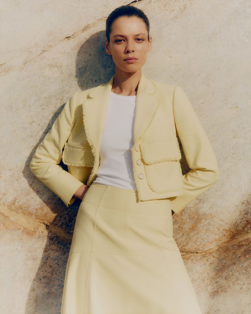Adult female poses wearing a light yellow skirt and jacket in front of a light-colored wall. The photo is shot at eye-level.