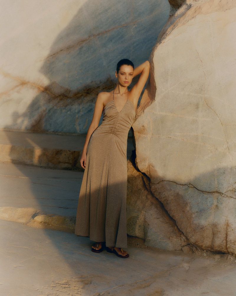 An adult woman poses in a halter dress, leaning against a rock in an outdoor setting. The lighting emphasizes shadows and textures.
