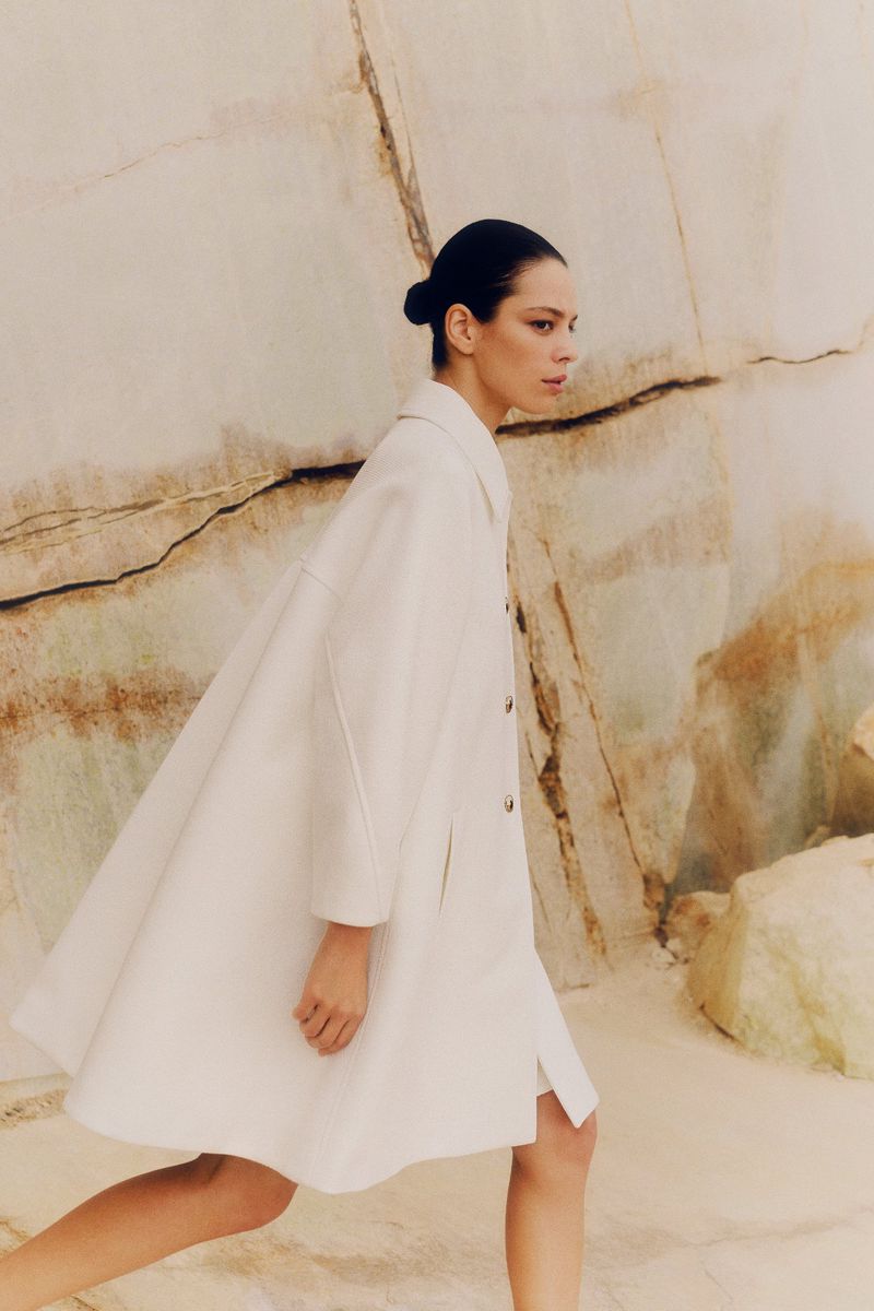 A woman in a white coat walks against a marble background, creating a minimalist fashion editorial with neutral tones and an elegant silhouette.