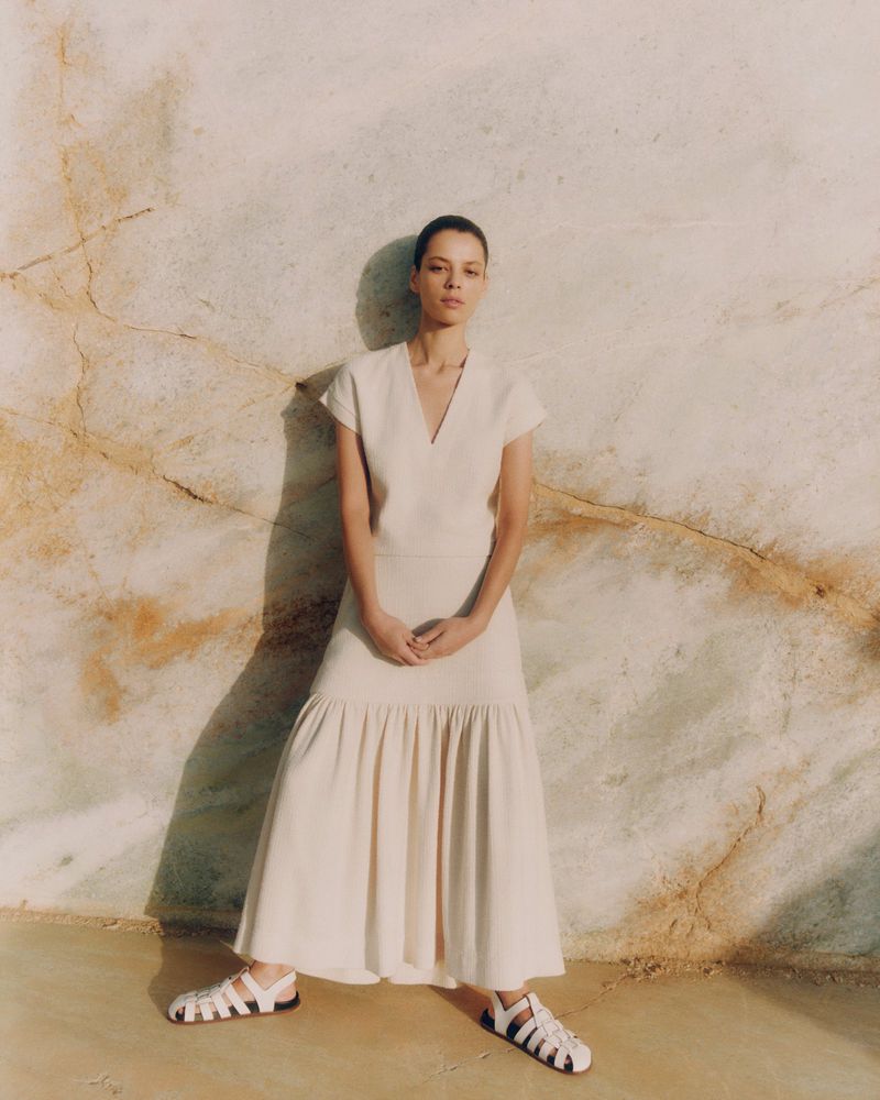 Elegant woman in a cream-colored dress and white strappy sandals poses against a textured marble wall, bathed in soft, diffused light.