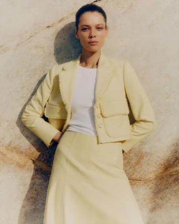 Model wearing a pale yellow tweed jacket and matching skirt leaning against a natural stone wall