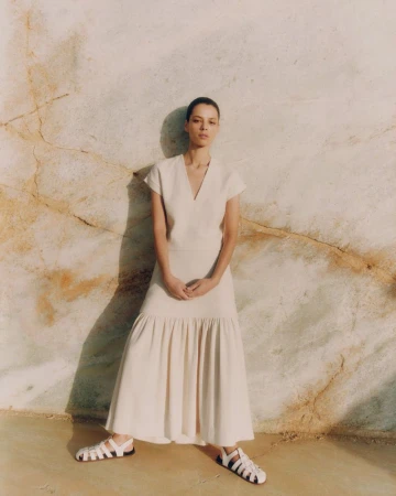 Model in a cream short-sleeve dress and white gladiator sandals standing against a textured stone wall