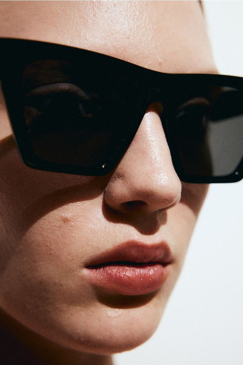 Close-up of a woman's face wearing black, square-framed sunglasses in bright, direct sunlight against a white background.