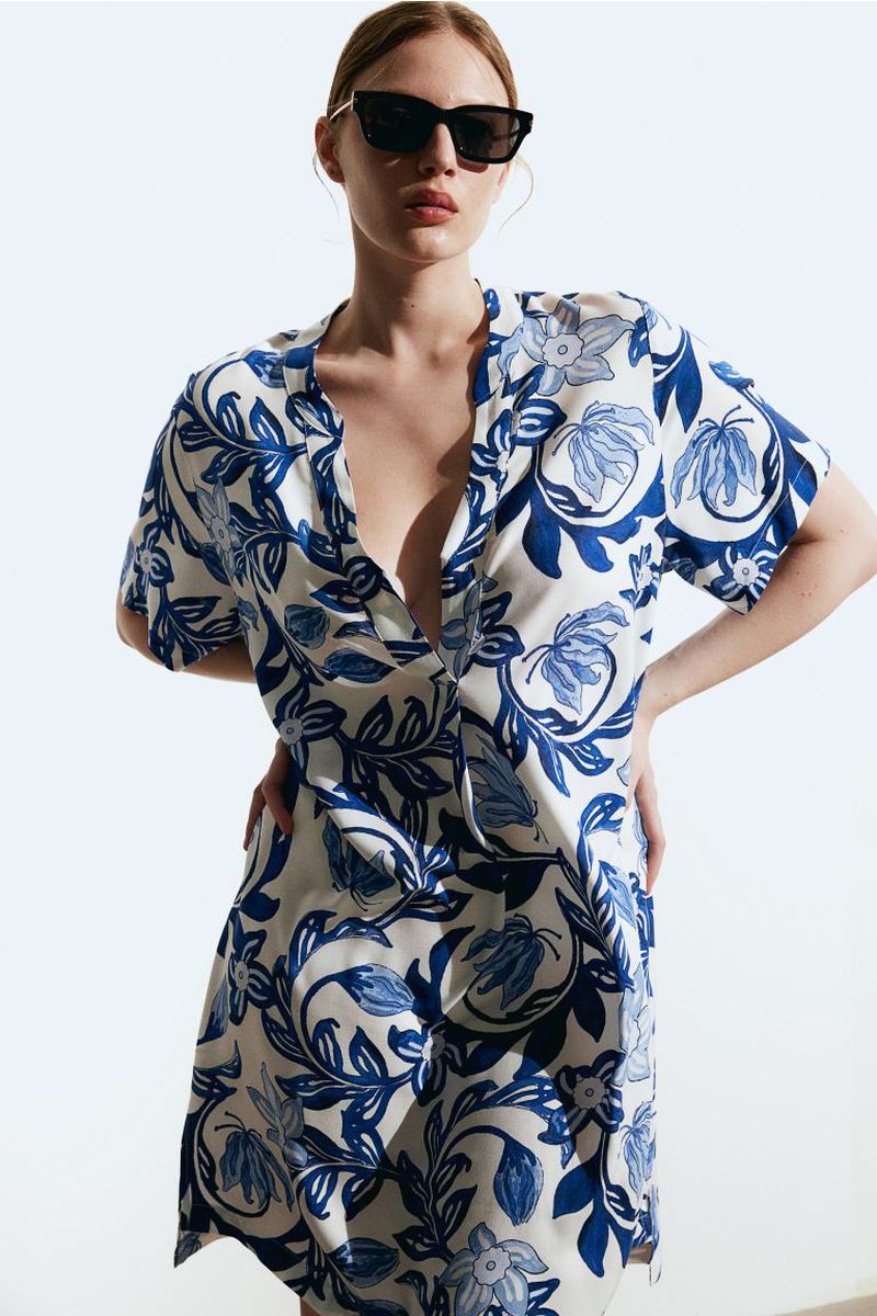 Model wearing dark sunglasses and a blue and white patterned dress against a bright white background