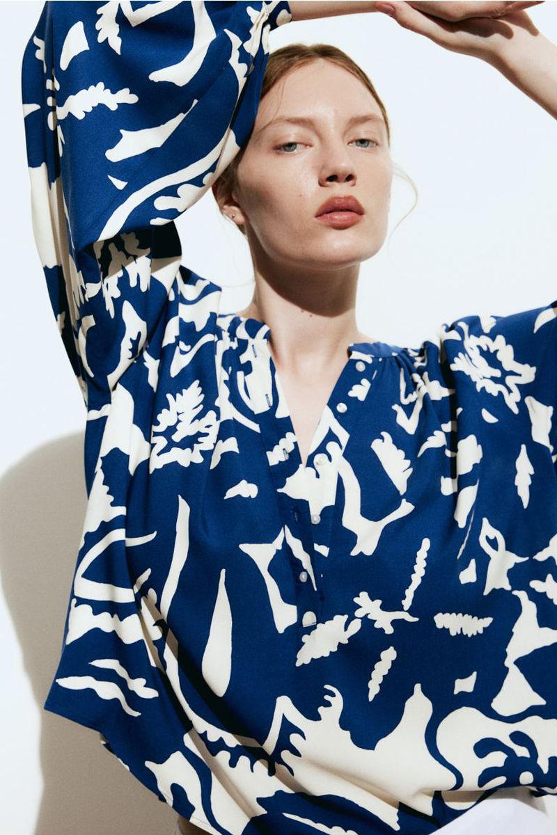 Close-up of a fair-skinned woman wearing a patterned, dark-blue and cream-colored V-neck blouse against a bright background.