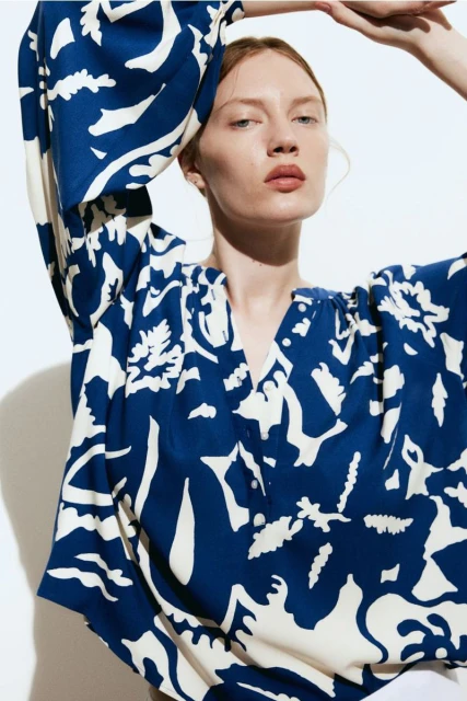 Close-up of a fair-skinned woman wearing a patterned, dark-blue and cream-colored V-neck blouse against a bright background.