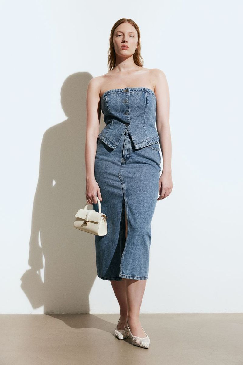 A woman wearing a denim tube top and midi skirt, holding a white purse and posing against a white background in a fashion shot.