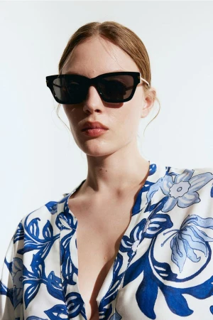 Model wearing dark sunglasses and a white and blue floral top against a plain light background