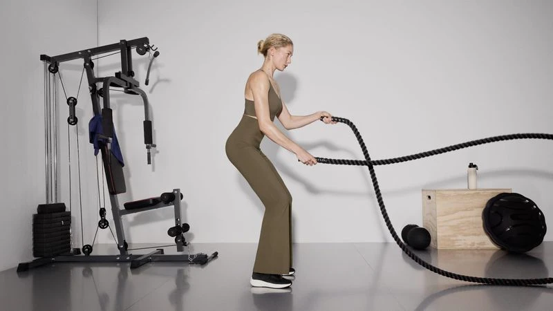 An adult female in sportswear uses battle ropes in an indoor setting with a multi-gym, weight plates, and a water bottle nearby.