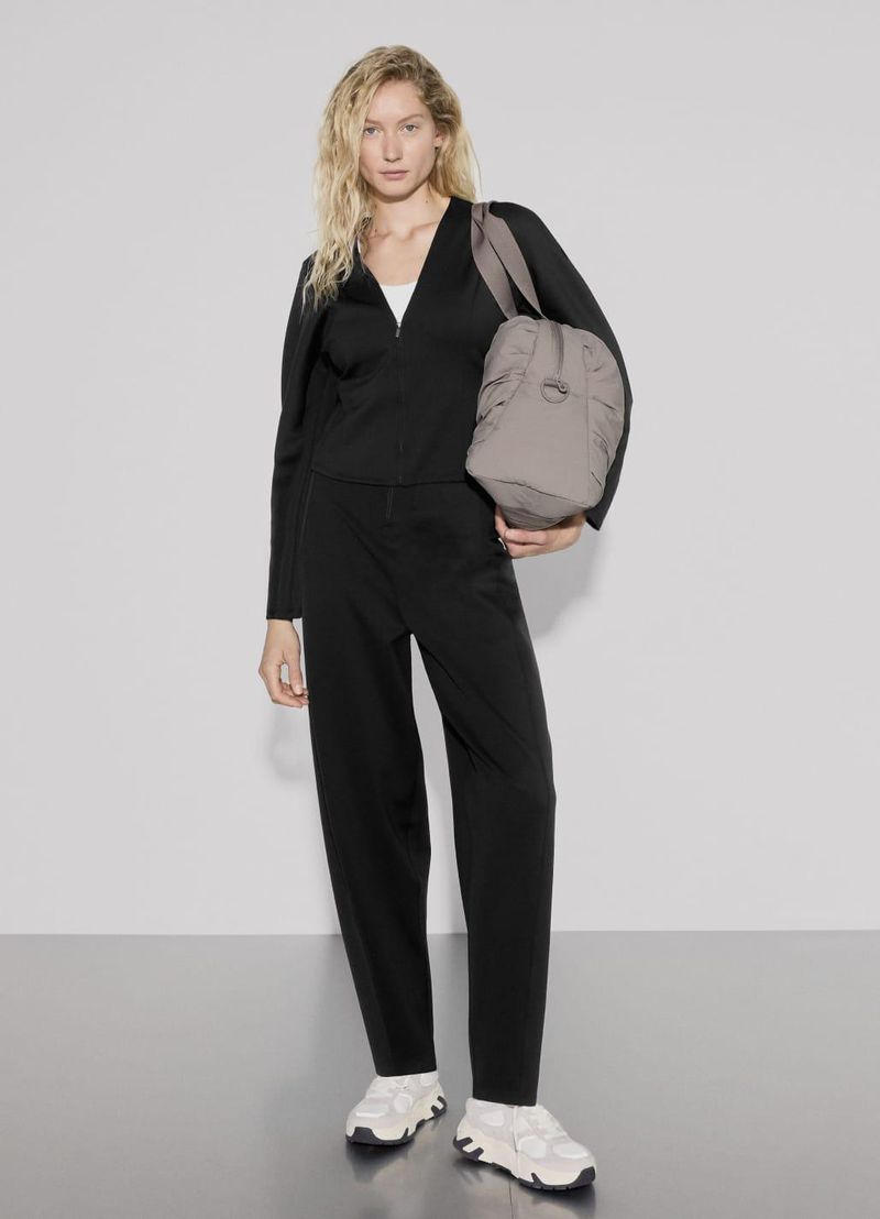 Blonde woman poses in black outfit with tote bag indoors against a white wall in a fashion context, full shot, lifestyle.