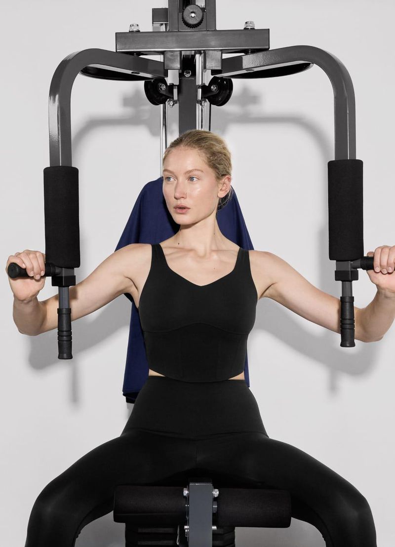 Woman using a black seated chest press machine in a brightly lit studio. The adult female is dressed in black workout clothes.