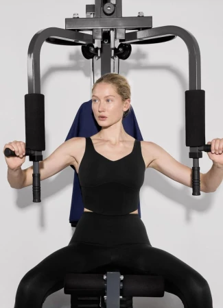 Woman using a black seated chest press machine in a brightly lit studio. The adult female is dressed in black workout clothes.