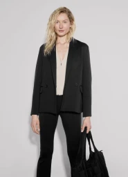 Model in black blazer and trousers holding a dark handbag against a plain grey studio backdrop