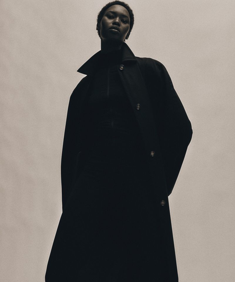 Fashionable adult woman poses in a long, dark overcoat against a plain background in a studio shot, creating a striking, moody image.