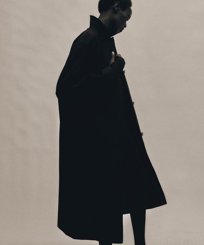 Fashion shot of a Black man wearing a long, dark overcoat in a photo studio with an off-white background, highlighting sleek lines and modern style.