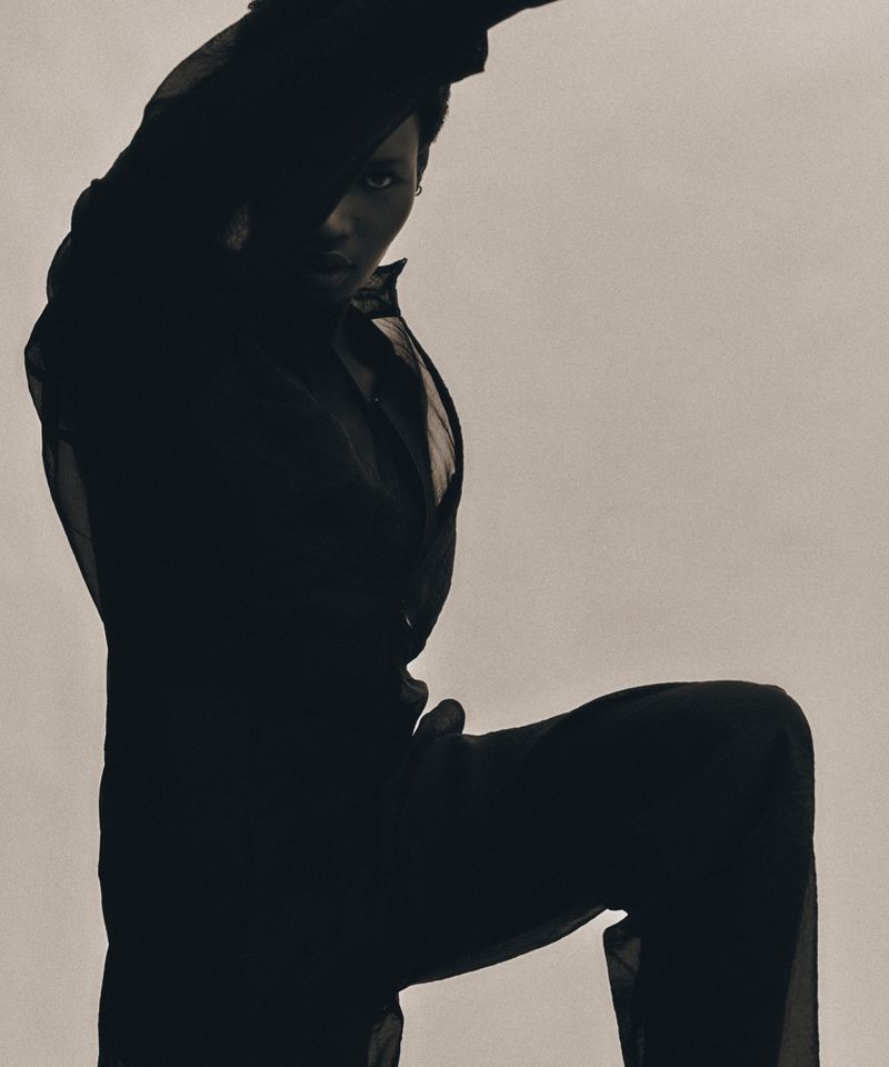 Striking full shot of a dark-skinned person in a sheer black outfit, posed against a light grey background, highlighted by studio lighting.