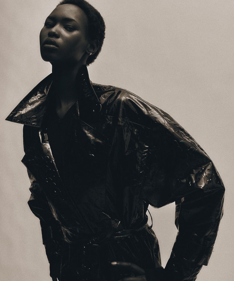 Elegant studio portrait of a young Black woman in a sleek black trench coat against a grey backdrop, fashion-forward and confident.