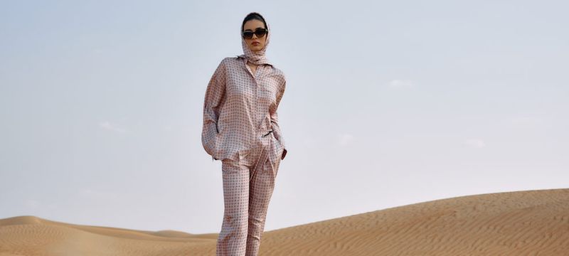 A woman in a patterned pajama set and headscarf stands in a desert under a clear sky, posing for a fashion shot with sand dunes.