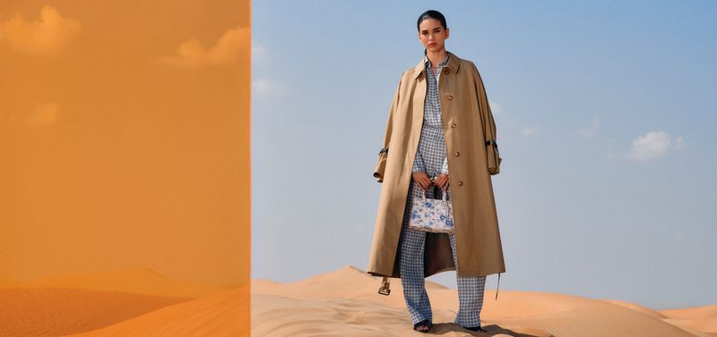 Model standing on sand dunes in a long camel trench coat and blue patterned trousers, holding a printed handbag