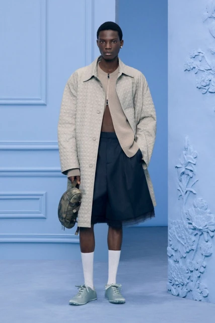 Fashionable man in an overcoat, shorts, and sneakers stands in a studio with a matching backdrop, accessorized with a small bag.