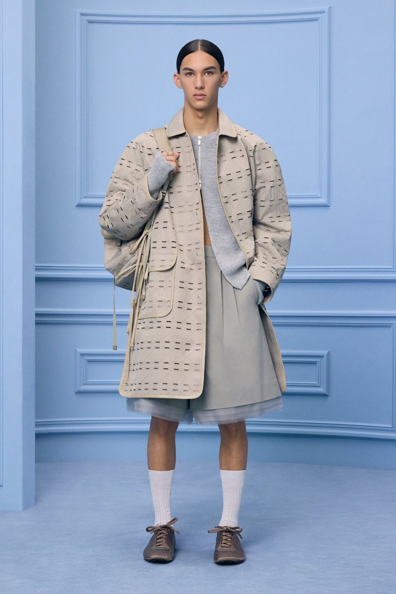 Male fashion model poses in neutral-tone coat, shorts, and sweater in a studio setting with a light blue decorative wall.