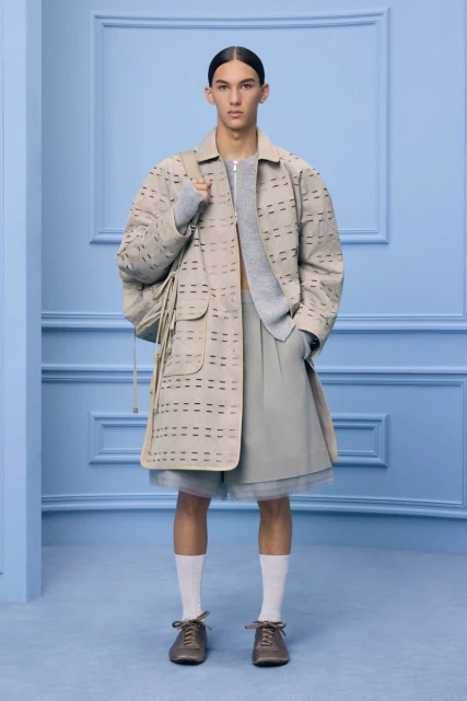 Male fashion model poses in neutral-tone coat, shorts, and sweater in a studio setting with a light blue decorative wall.