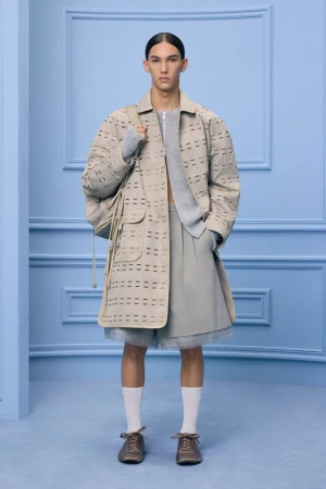 Male fashion model poses in neutral-tone coat, shorts, and sweater in a studio setting with a light blue decorative wall.