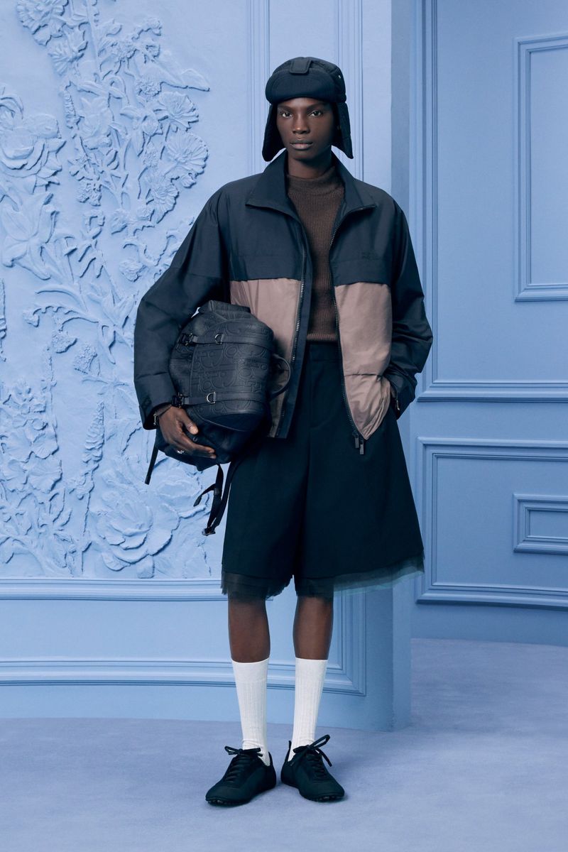 Dark-skinned male model in a color-blocked jacket, culottes, socks, and sneakers, holding a textured backpack, in a blue-toned studio.