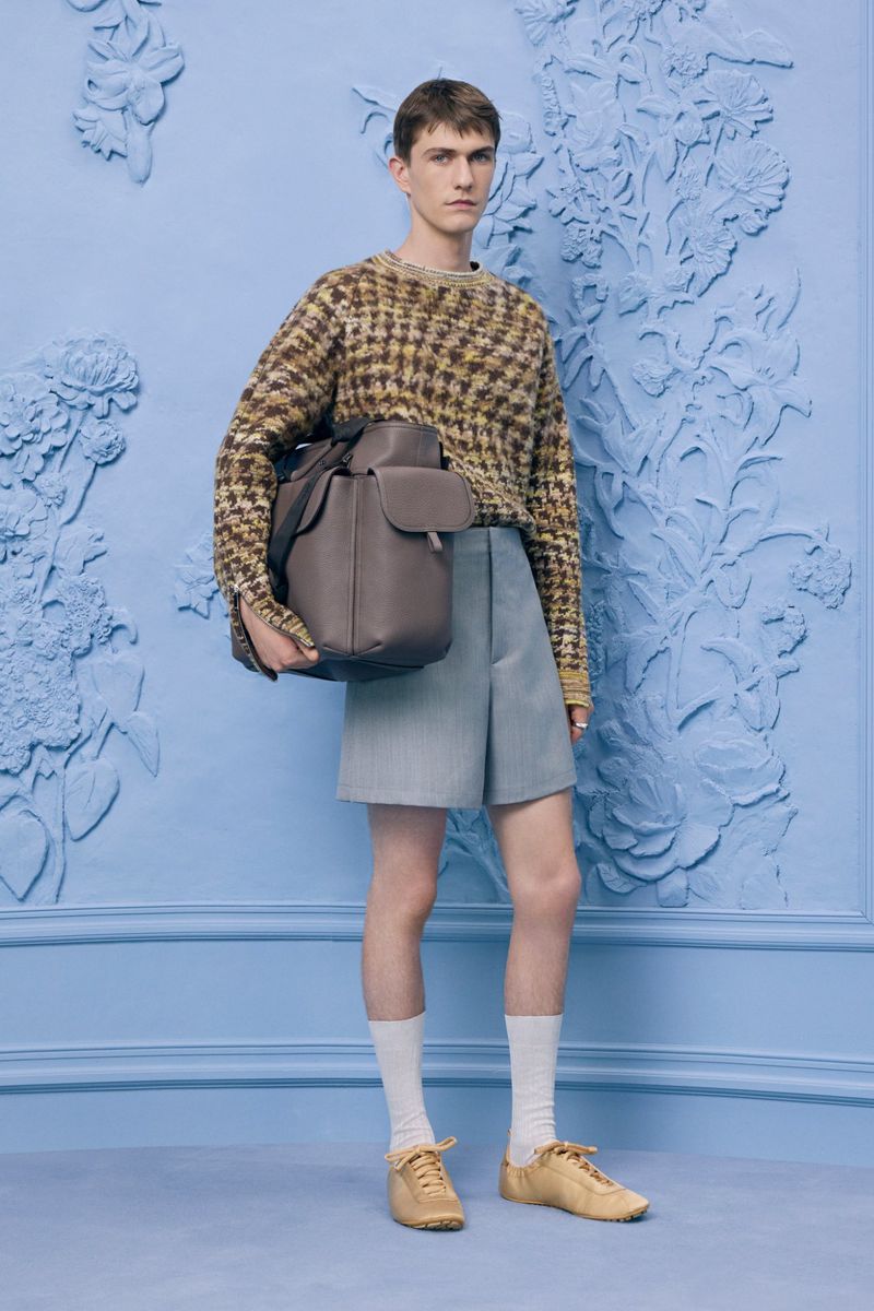A young adult male poses in a patterned sweater, light blue shorts, socks, and shoes with a leather backpack against a blue floral backdrop.
