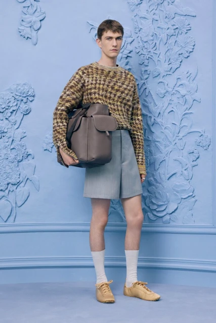 A young adult male poses in a patterned sweater, light blue shorts, socks, and shoes with a leather backpack against a blue floral backdrop.