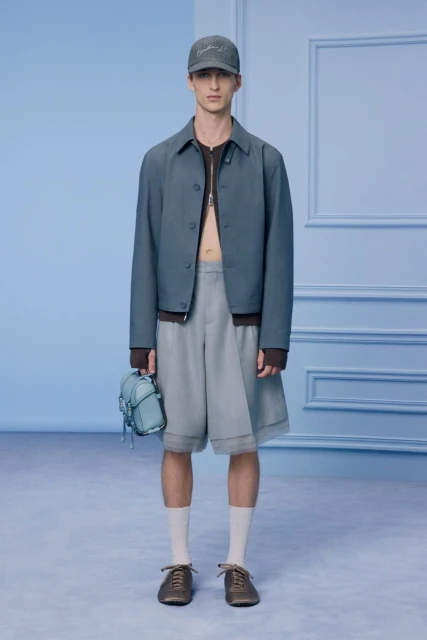 Fashion shot of a model in a blue jacket, grey shorts, socks, and brown shoes in a studio setting with soft, diffused lighting.