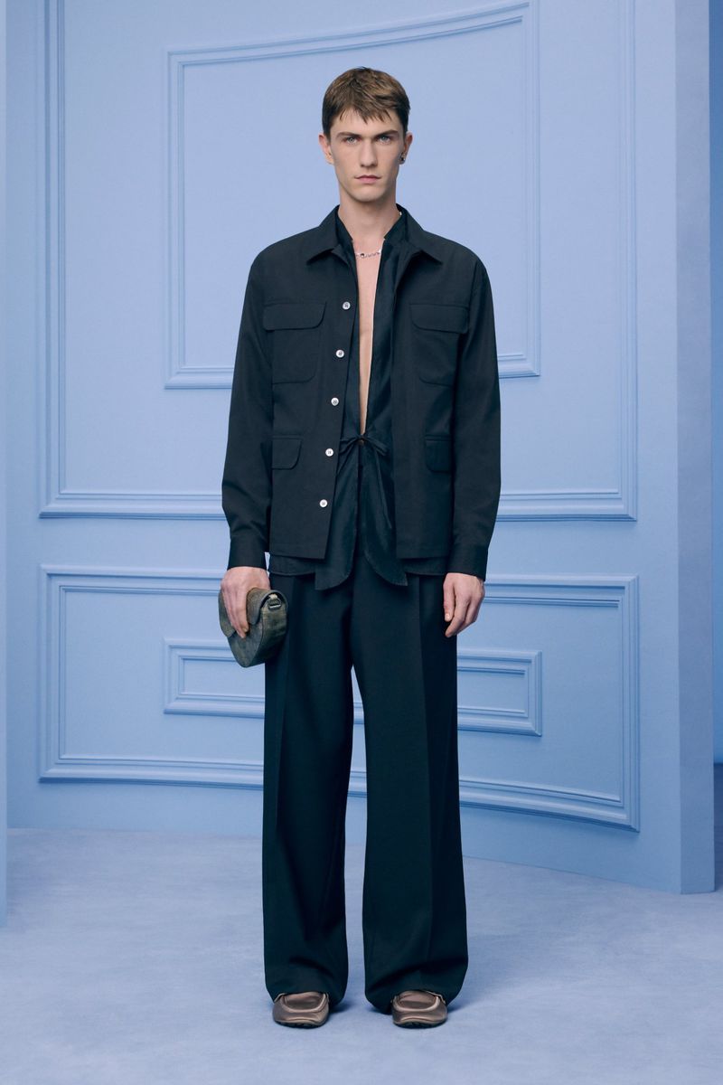 A young adult male stands in a full-length studio shot, wearing a dark open shirt, jacket, dark wide-legged pants, and brown loafers, carrying a clutch.