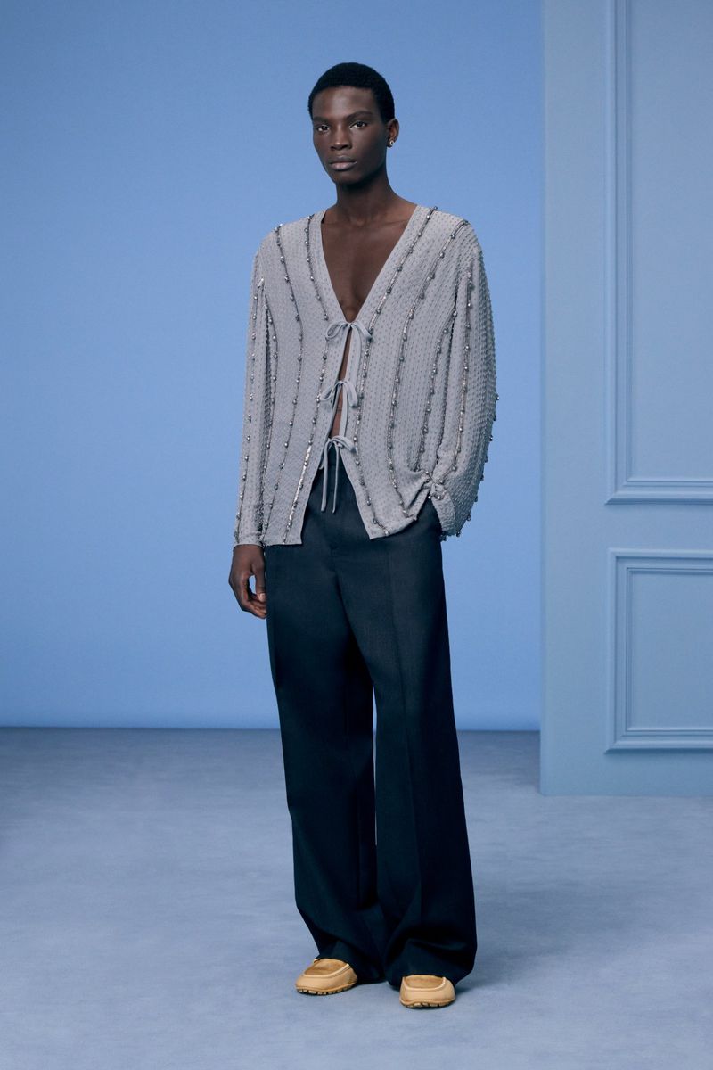 An African-American man poses in a light grey top with black pants indoors. He wears light brown shoes and stands against a blue wall.