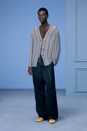 An African-American man poses in a light grey top with black pants indoors. He wears light brown shoes and stands against a blue wall.