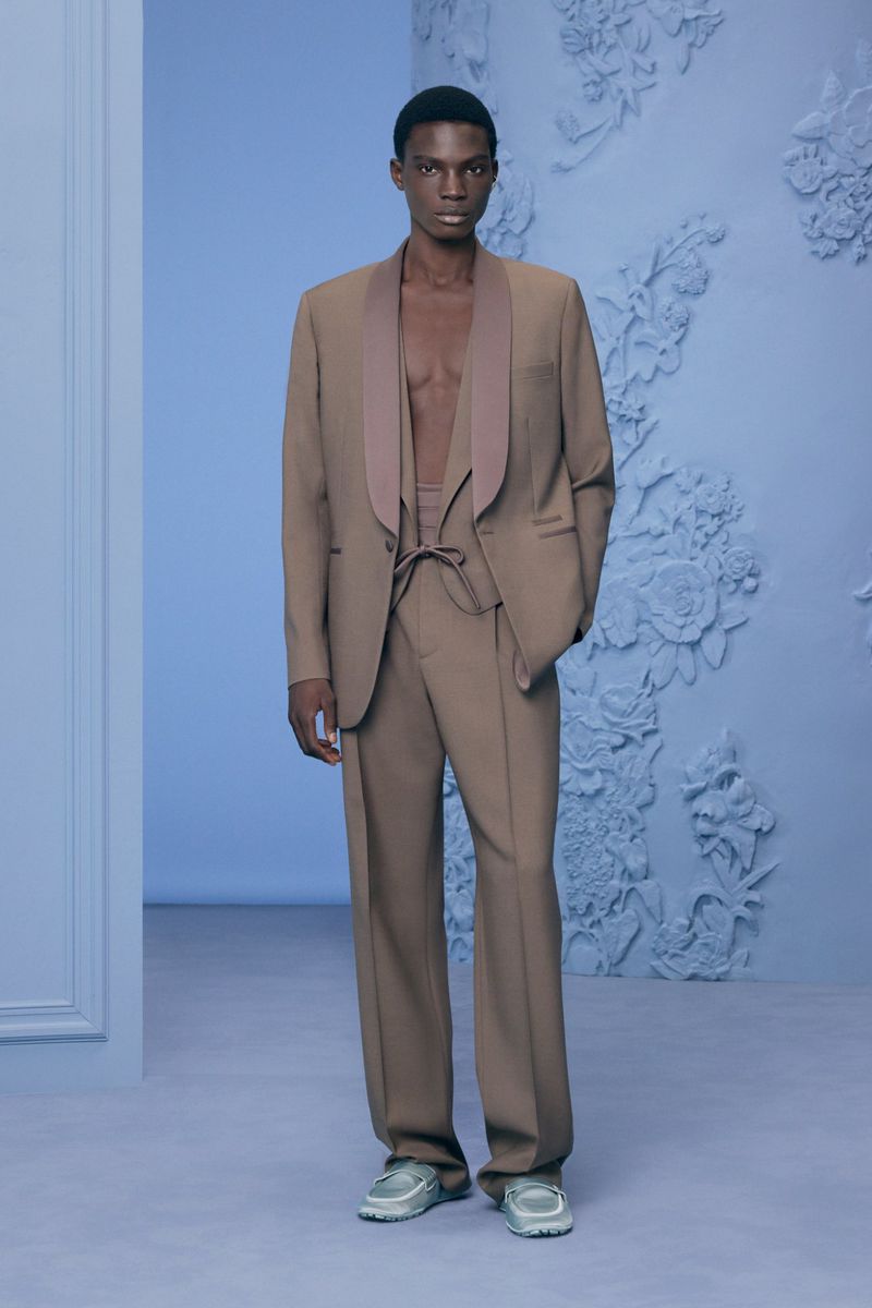 Fashionable African-American man wearing a tan suit and sneakers in front of a light blue textured wall, posing in a photo studio.