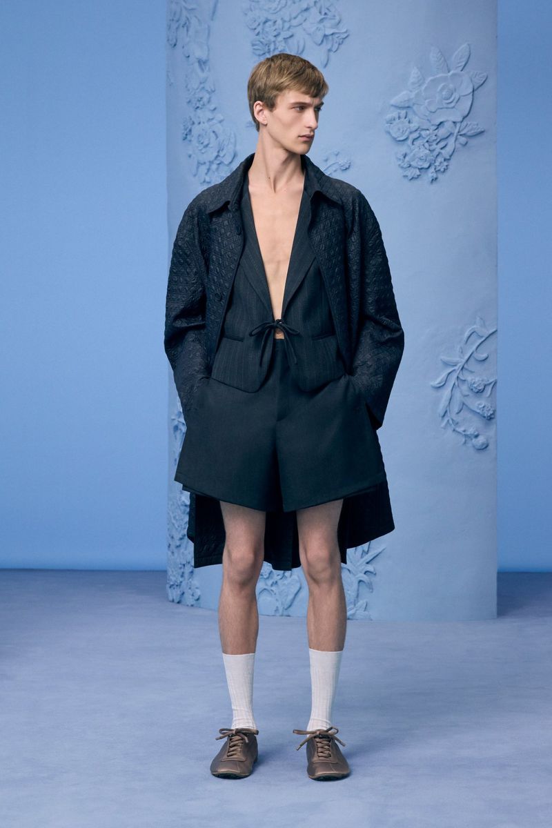Male model standing in a dark navy tailored shorts and jacket set with white socks against a powder blue studio background.