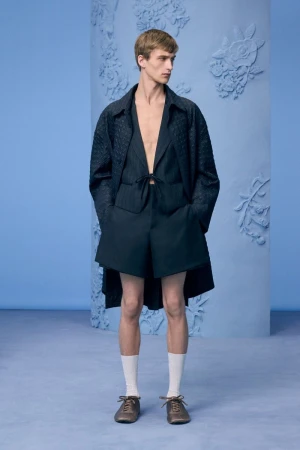 Male model standing in a dark navy tailored shorts and jacket set with white socks against a powder blue studio background.