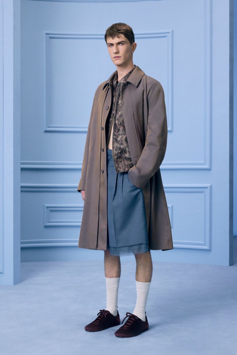 Full-length portrait of a male model wearing a brown overcoat, patterned shirt, blue shorts, white socks, and dark shoes against a blue backdrop.