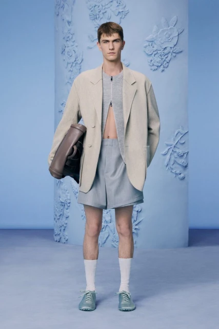 Full-length shot of a man in a beige blazer, gray shorts, and white socks, holding a large brown leather bag in a light blue studio.