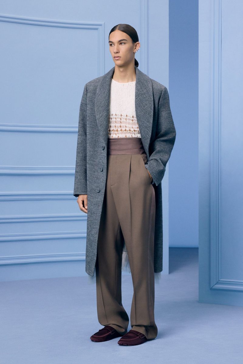 A slender man wearing a grey overcoat, beige sweater, and brown trousers poses in a light blue room with simple wall moldings.