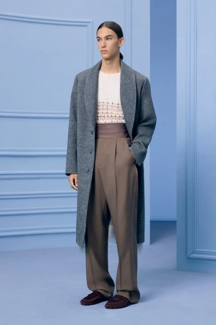 A slender man wearing a grey overcoat, beige sweater, and brown trousers poses in a light blue room with simple wall moldings.