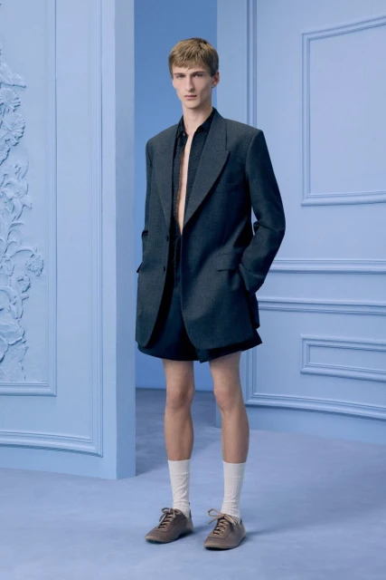 An adult male model poses in a navy blue blazer and shorts, paired with socks and brown shoes against a soft blue studio backdrop.
