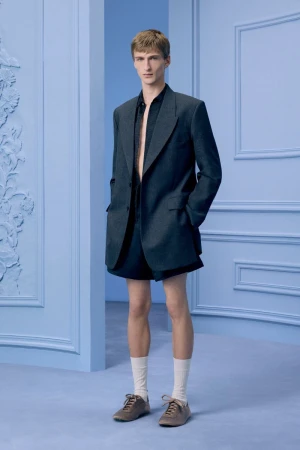 An adult male model poses in a navy blue blazer and shorts, paired with socks and brown shoes against a soft blue studio backdrop.
