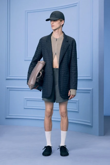 Full-length studio shot of a man in a black blazer, shorts, socks, and black baseball cap holding a grey bag in a blue studio.